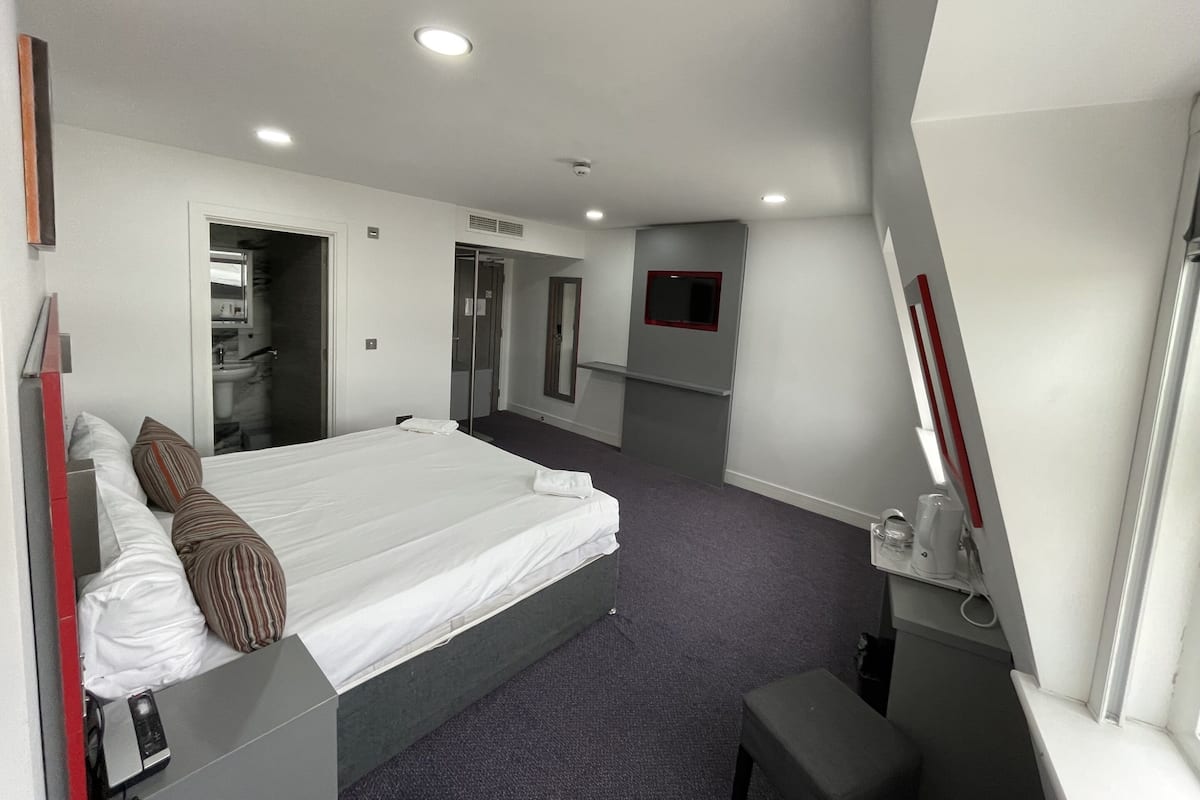 A modern hotel room with a large bed, a desk, a chair, a mirror, and a small table with a coffee maker.