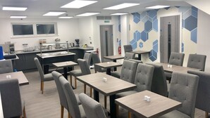Un comedor de hotel moderno con múltiples mesas y sillas, una zona de cocina con máquinas de café, y una sala de conferencias con un diseño geométrico en la pared.