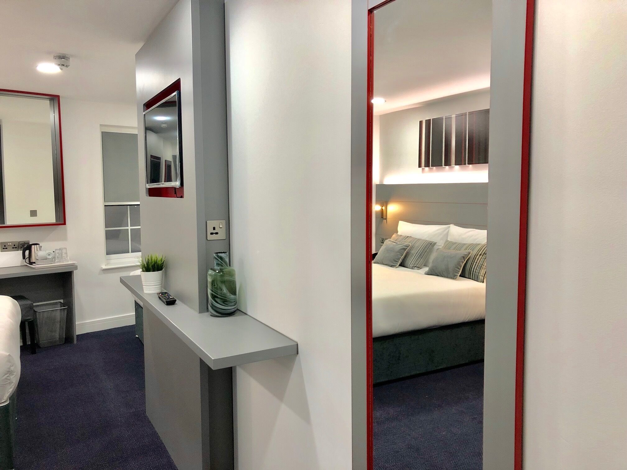 Une chambre d’hôtel moderne avec un grand miroir, une table de chevet ornée d’un vase, un lit avec des oreillers et une petite fenêtre.