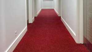 Un pasillo con alfombra roja, paredes blancas y un ventanal al final.