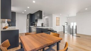 A modern kitchen with dark cabinetry, a wooden dining table, and a view of the living area. - The Rodney House - Midcentury, Modern Sleeps 8 (Reno)