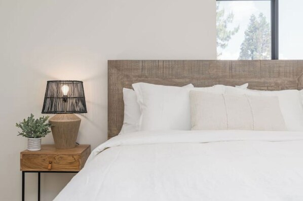 A neatly made bed with white bedding, a wooden nightstand with a woven lamp, and a potted plant.