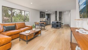 A modern living room with a leather sofa, a wooden coffee table, and a dining area with a kitchen in the background. - The Rodney House - Midcentury, Modern Sleeps 8 (Reno)