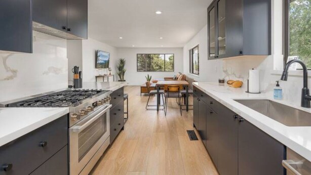 A modern kitchen with dark cabinetry, a central island, and a dining area with a view of the outdoors. - The Rodney House - Midcentury, Modern Sleeps 8 (Reno)