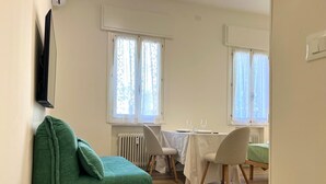Habitació amb un sofà verd, una taula de dinar amb caires i dues finestres amb cortines.