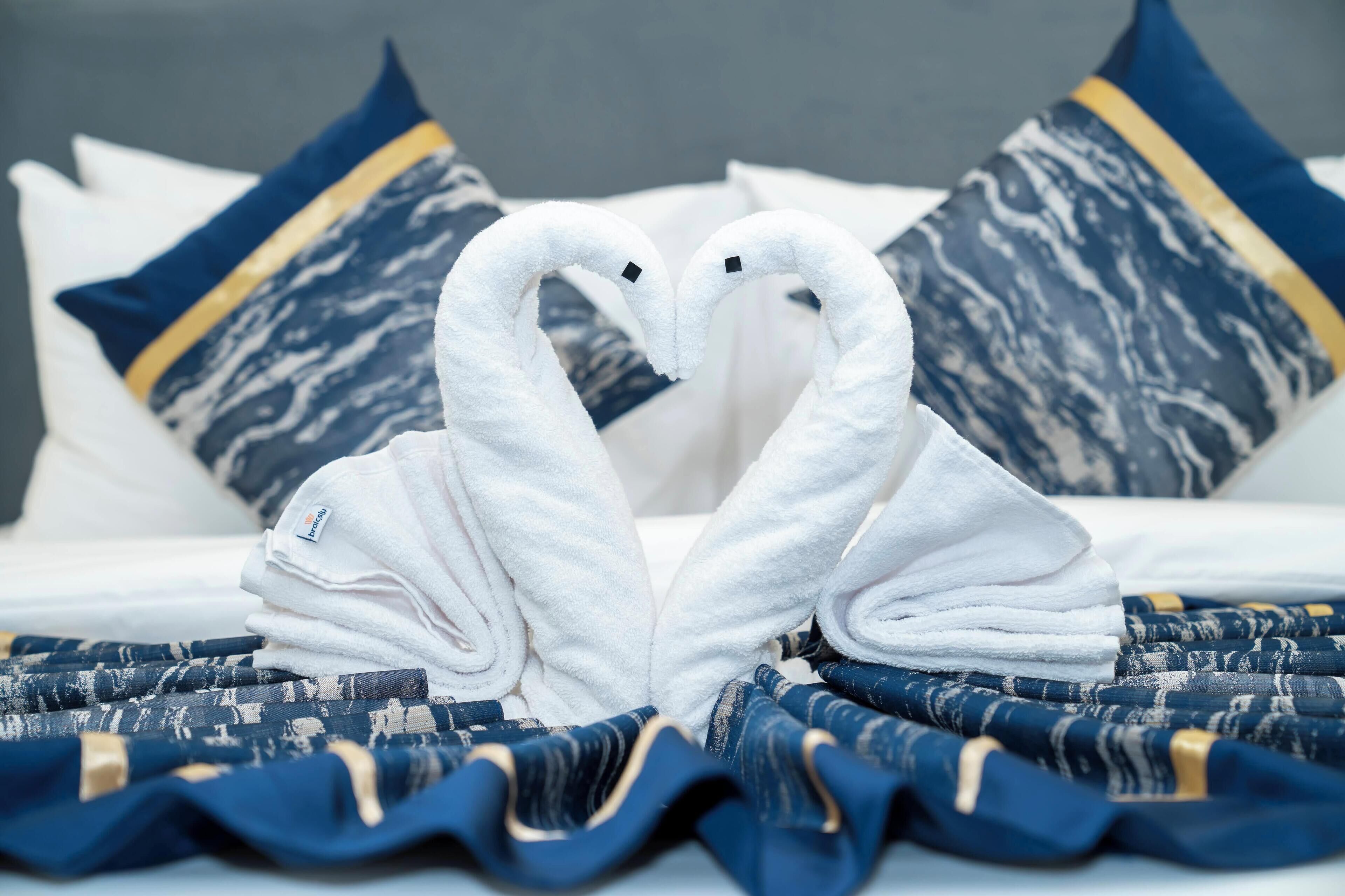 Una cama con almohadillas de patrón blanco y azul, una manta a rayas azul y blanco, y dos toallas blancas dispuestas en forma de corazón.
