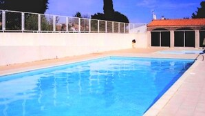 En swimmingpool med et hvidt hegn, et overdækket område med vinduer og en bygning med rødt tag i baggrunden.