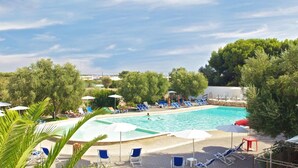 Una piscina con sillas de descanso y sombrillas, rodeada de árboles y un cielo despejado.
