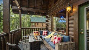 A cozy log cabin with a wooden deck, a sofa with plaid cushions, a table with drinks, and a neon sign reading "The American."