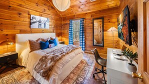 A cozy log cabin bedroom with a large bed, a desk, and a window with curtains.