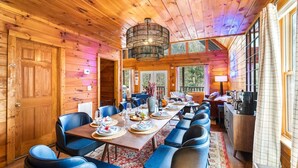 A wooden cabin with a dining area featuring a long table, blue chairs, and a chandelier.