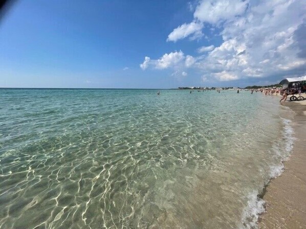 Una playa con aguas poco profundas y transparentes, orilla arenosa y algunas personas a lo lejos.