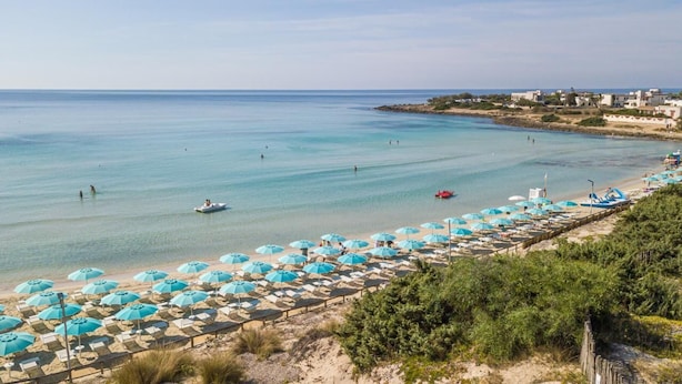 Une plage bordée de rangées de parasols et de chaises longues, une mer d’un bleu limpide et quelques bateaux.