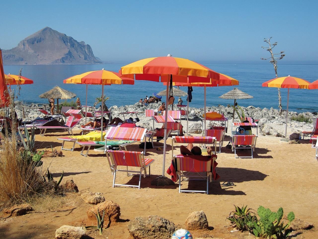 A beach with multiple sun loungers and umbrellas, rocky terrain, and a mountain in the background.