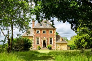 Une maison traditionnelle en pierre, dotée d’une entrée centrale et entourée d’une végétation luxuriante et d’arbres.