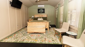 A bedroom with a bed, a chair, a small table, a ceiling fan, a TV mounted on the wall, and a window with curtains.