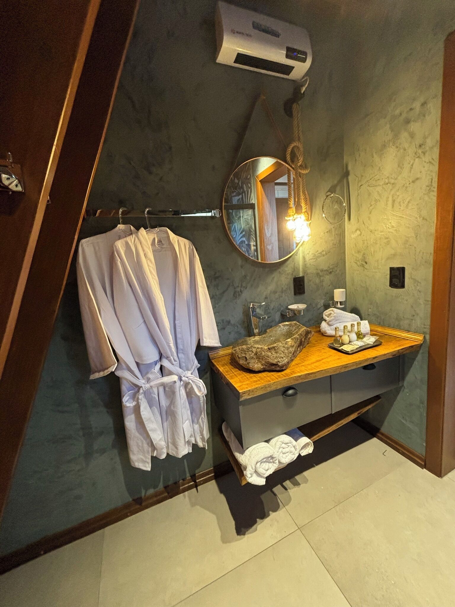 A bathroom with a wooden vanity, a round mirror, a robe hanging on a rack, and a towel on the floor.