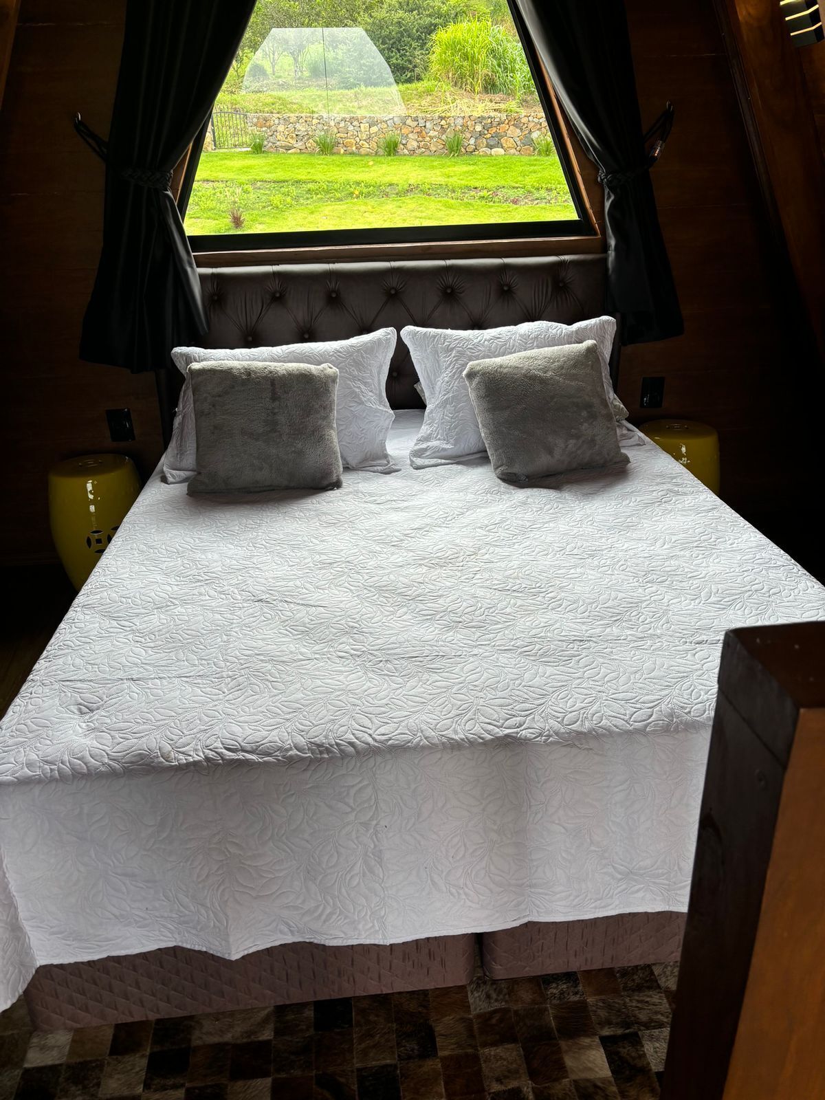 A bed with a white quilted bedspread and two pillows, a window with a view of a garden, and a wooden headboard.