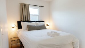 Un lit bien fait, avec du linge de lit blanc et une serviette pliée, une petite table ronde à côté, et deux lampes fixées au mur.