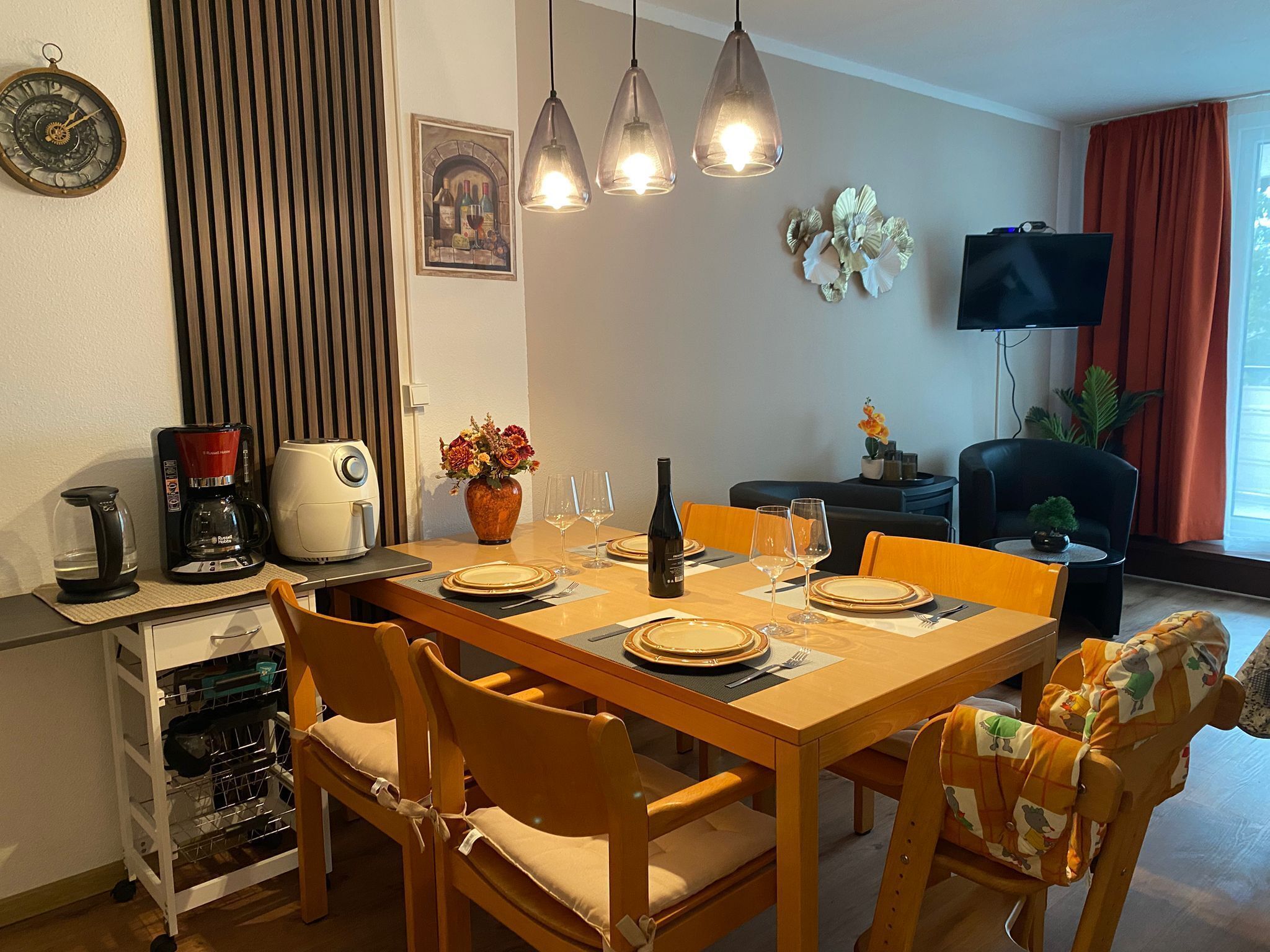 En matplats med ett träbord för fyra personer, stolar, en kaffemaskin, en tv och ett fönster med gardiner.