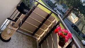 A balcony with a view of a street, a hanging plant, a wicker chair, and a table with cushions. - 2 room family apartment "Vier Jahreszeiten" (Sankt Englmar)