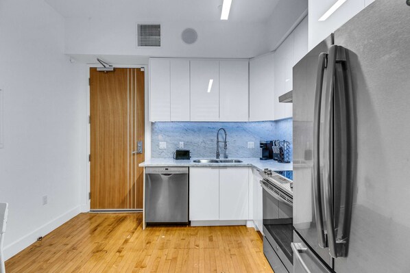 A modern kitchen with white cabinets, a stainless steel refrigerator, a dishwasher, and a wooden door.