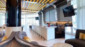 Un hall d’hôtel moderne doté d’un bar central, de différents espaces aménagés pour s’asseoir et d’un grand écran de télévision.