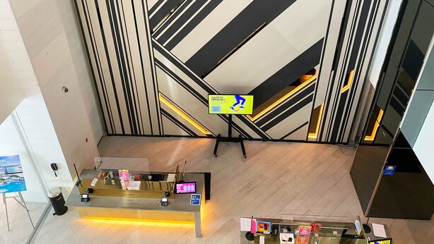 A modern reception area with a digital screen displaying safety instructions, a reception desk, and a staircase with a striped wall design. - 5-star Condo at The W Residences 1BR by Oneluxstay (Fort Lauderdale)