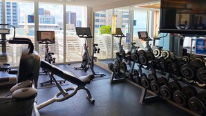 Une salle de sport moderne équipée de divers appareils d’exercice, de grandes fenêtres offrant une vue sur la ville et d’un écran de télévision.