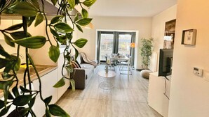 A modern living room with a sofa, coffee table, and a dining area with chairs. There is a large window with curtains, a plant, and a television on the wall. - Cozy 1BR City Centre Near Meir Shopping by Oneluxstay (Antwerp)