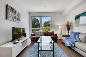 A modern living room with a glass coffee table, a brown leather sofa, a white entertainment unit, and a large window offering a view of trees.