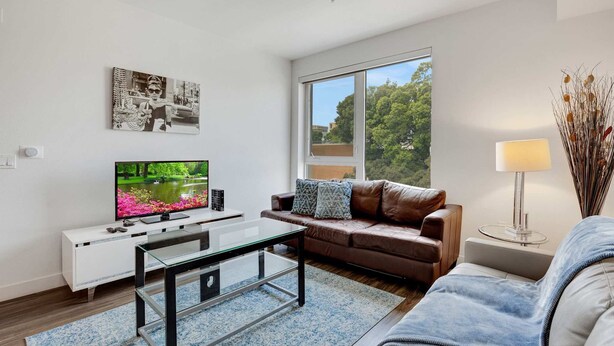 A modern living room with a flat-screen TV, a glass coffee table, a brown leather sofa, a white console table, and a large window offering a view of trees. - Stylish 1BR Family Apartment by Dtla (Los Angeles)