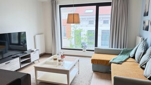 A modern living room with a flat-screen TV, a coffee table, a sofa, and a large window offering a city view. - Spacious 3BR 2/5 BA Near Diamond District by Oneluxstay (Antwerp)