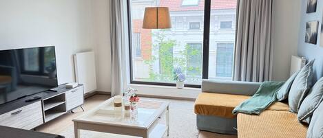 A modern living room with a flat-screen TV, a coffee table, a sofa, and a large window offering a city view.