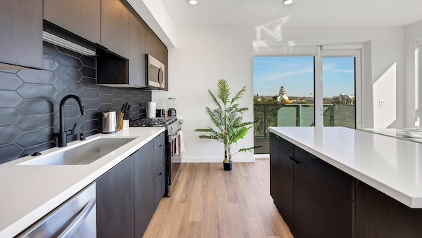 A modern kitchen with dark cabinetry, a white countertop, and a view of the city through large windows. - Luxury 2BR Hollywood Pool View by Oneluxstay (Los Angeles)