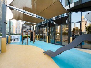 Un parque infantil interior con estructuras para escalar y un tobogán, rodeado de paredes de vidrio con vistas a la ciudad.