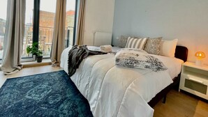Un dormitorio con una cama grande, una mesita de noche, una lámpara, una ventana con cortinas, una planta en maceta y vistas de edificios al exterior.