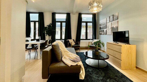 Una sala moderna con un sofá, una mesa de centro y un televisor. Hay ventanas amplias con cortinas, una zona de comedor y una planta en el alféizar.
