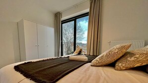Una cama bien hecha con un edredón oscuro, una ventana con cortinas y un armario blanco.