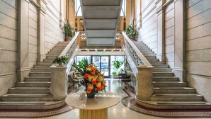A grand lobby with a marble staircase, a central round table with a floral arrangement, and a view of the entrance.