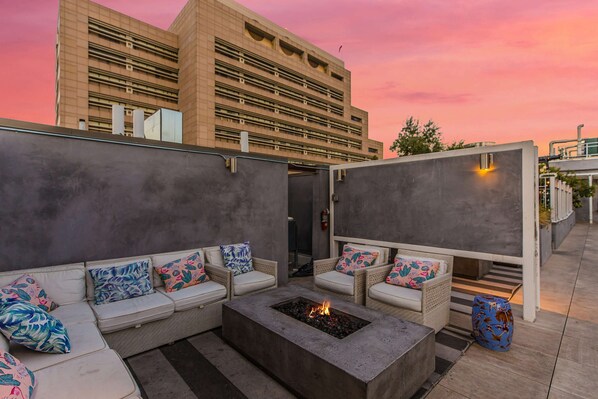 An outdoor seating area with sofas and a fire pit, set against a building with a pink sky.