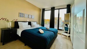 Ett hotellrum med en stor säng, ett nattduksbord, en vas med blommor och utsikt över staden genom stora fönster.