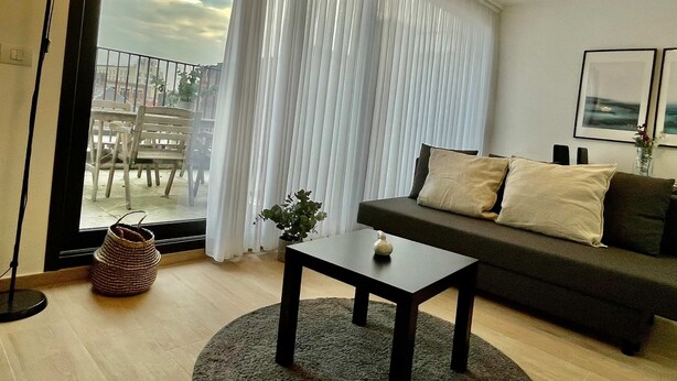 A modern living room with a dark sofa, a small table, and a large window with sheer curtains. - Modern 1BR Near Diamond District by Oneluxstay (Antwerp)