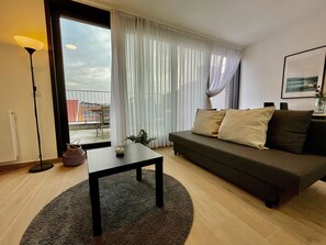 Sala de estar con sofá, una mesa y una lámpara. Hay una ventana con cortinas y vistas al exterior.