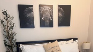 Una cama bien hecha con una manta gris, almohadas blancas y una almohada con el texto 'always stay humble & kind'. Hay tres cuadros enmarcados en la pared sobre la cama.