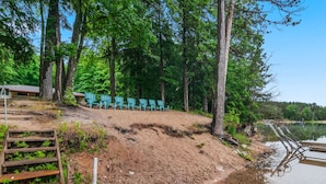 Zona junto a un lago con una escalera de madera que baja a una playa de arena, rodeada de árboles y una fila de sillas verdes.