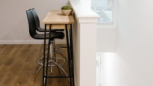 Een kleine kamer met witte wanden, een schuin dak, een houten tafel met twee zwarte stoelen en een raam met luiken.