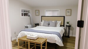 Een slaapkamer met een bed, nachtkastjes, een bankje, een airconditioning en een raam met jaloezieën.