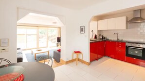 A modern kitchen with red cabinets, a black countertop, and a stainless steel range hood. There is a dining area with a glass table and chairs, and a small stool. - 4guests-2br-privategarden-freeparking (Aviemore)