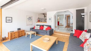 A modern living room with a grey sofa, a wooden coffee table, and a blue rug. There is a wooden cabinet and a small table with a vase. The room has white walls and a ceiling with exposed beams. - 4guests-2br-privategarden-freeparking (Aviemore)
